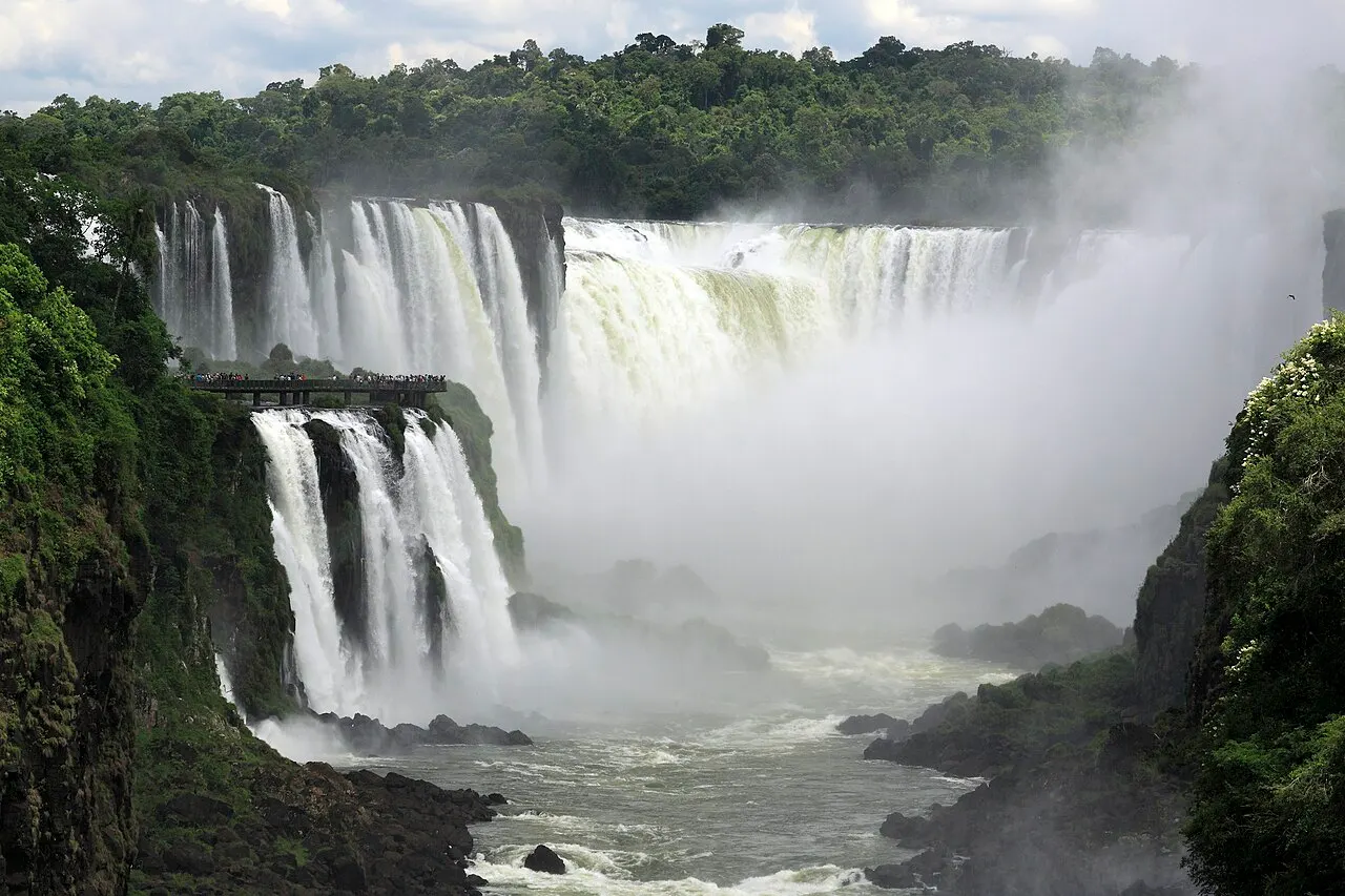 Uma passarela metálica com pessoas apoiada sobre a mata, diante de uma grande cortina de quedas d'água que desce em meio a uma densa nuvem de névoa, floresta ao redor sob céu nublado.
