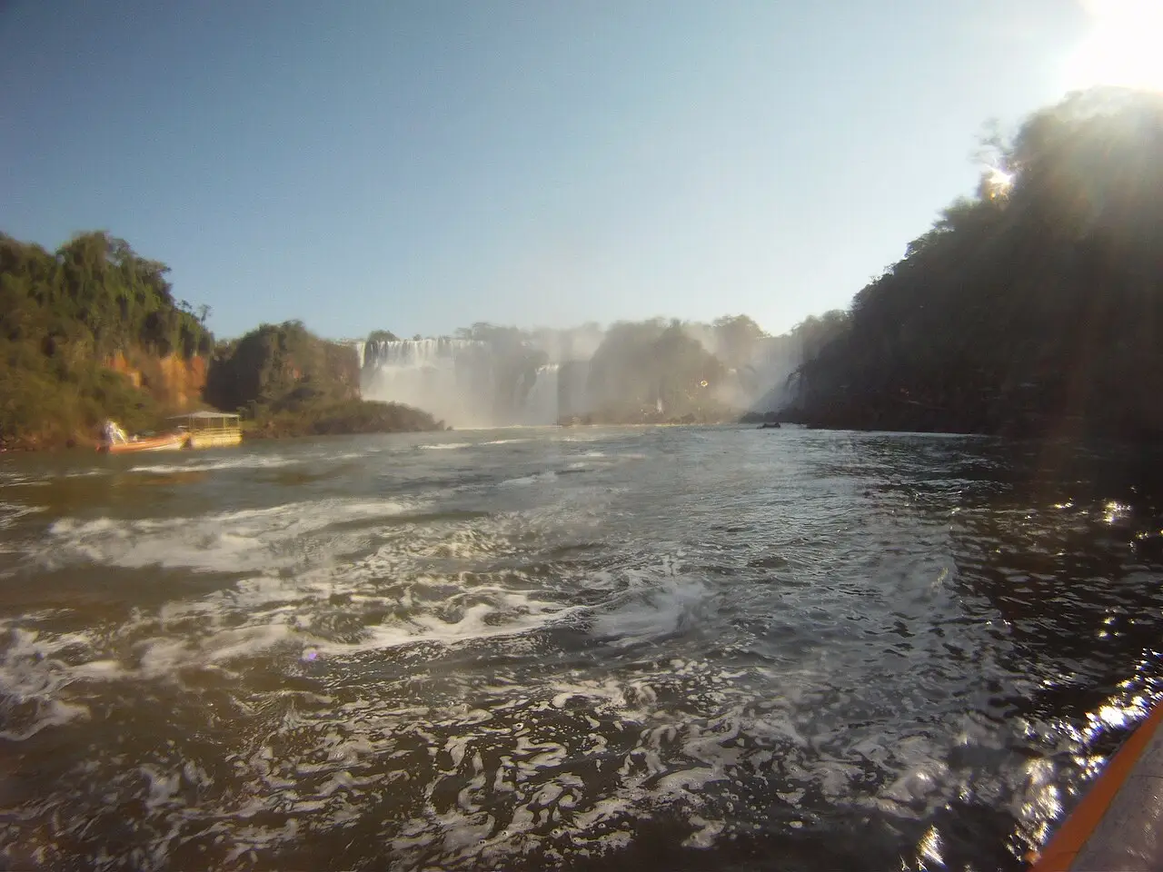 Cataratas ao fundo do rio, vistas da água no Macuco Safari, com um barco amarelo à esquerda e a borda do barco em primeiro plano.