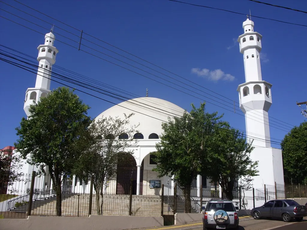 A mesquita branca tem cúpula e dois minaretes, com árvores e uma grade na frente, fios no céu e carros na rua.