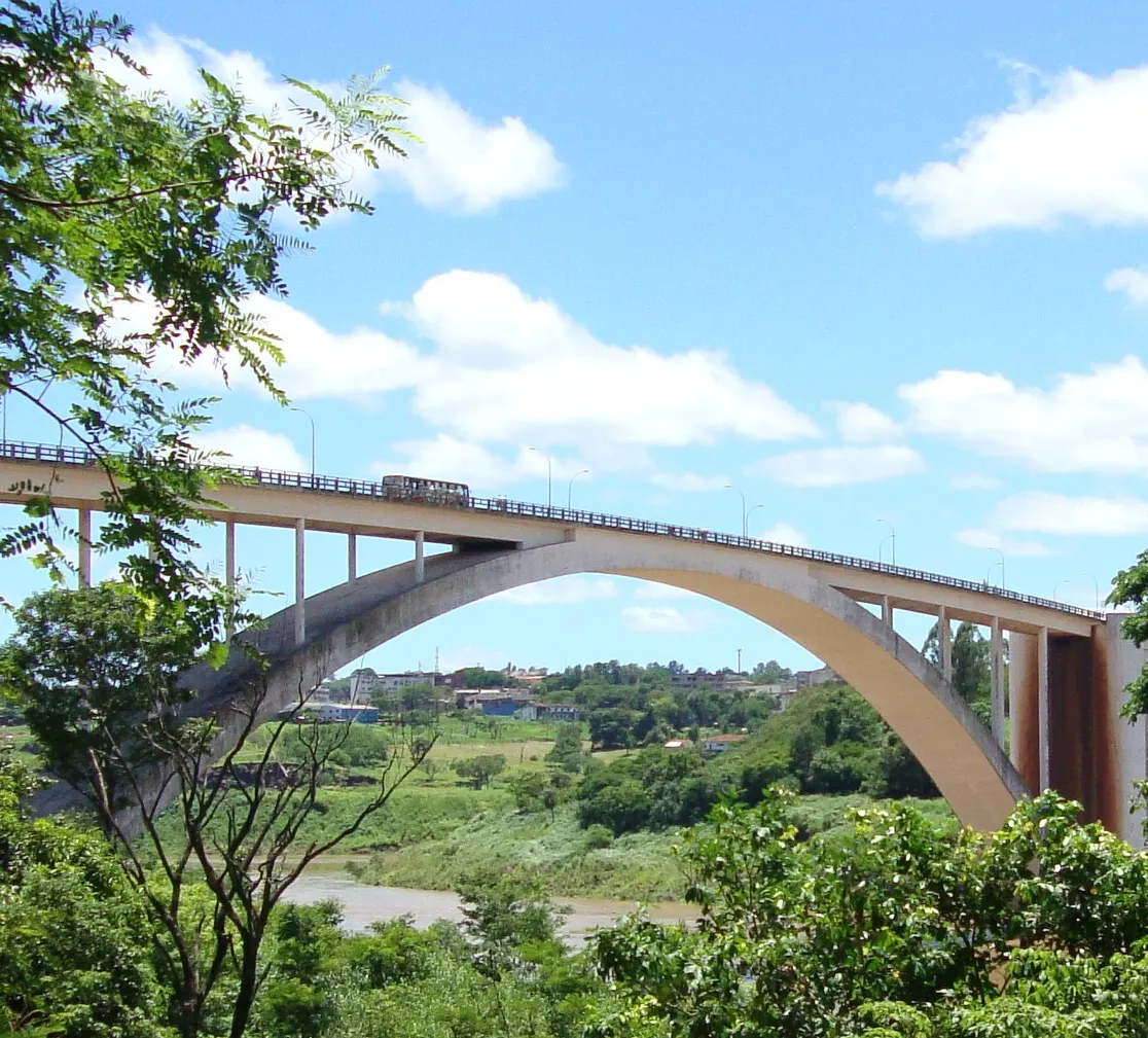 Um arco alto de concreto cruza o rio, com um veículo no tabuleiro, mata verde em volta e céu azul com nuvens.