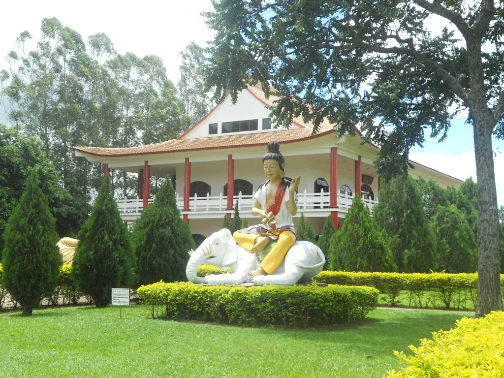 O prédio branco tem telhado alaranjado de beiral curvo e colunas vermelhas, e no jardim uma figura de manto amarelo está sobre uma escultura branca, entre arbustos.