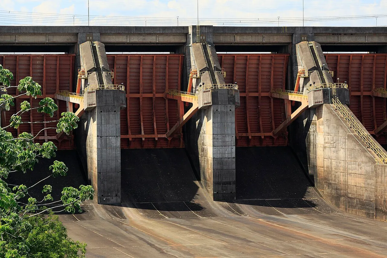 Três comportas vermelhas fechadas do vertedouro de Itaipu entre pilares de concreto, com escadas metálicas amarelas e o canal de descarga seco embaixo.