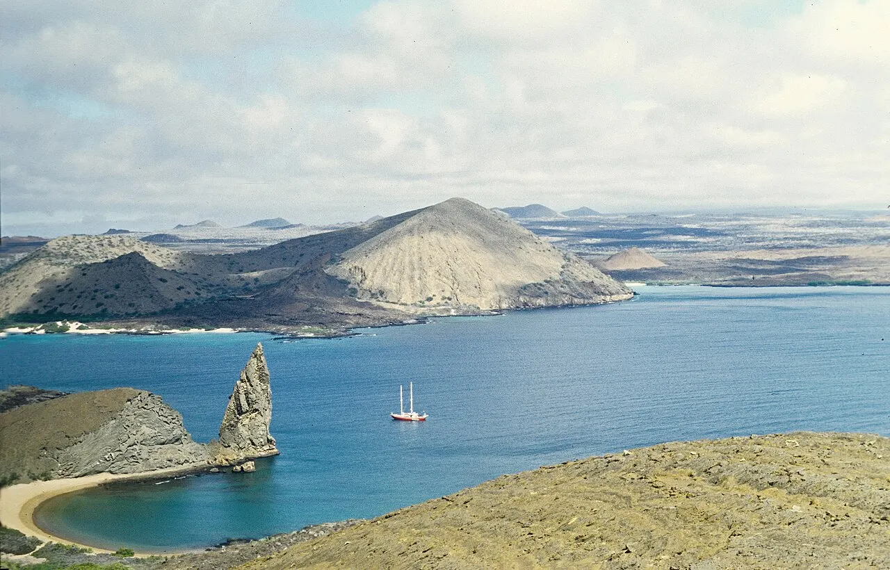 O obelisco de rocha do Pinnacle Rock na ponta de Bartolomé, com uma praia em meia-lua de areia clara à esquerda, um veleiro fundeado na baía azul e cones vulcânicos castanhos ao fundo.