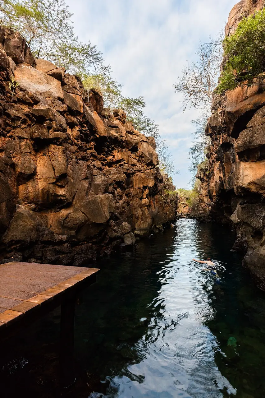 Passagem de água entre paredes rochosas, com uma plataforma de madeira à esquerda e uma pessoa nadando de costas.