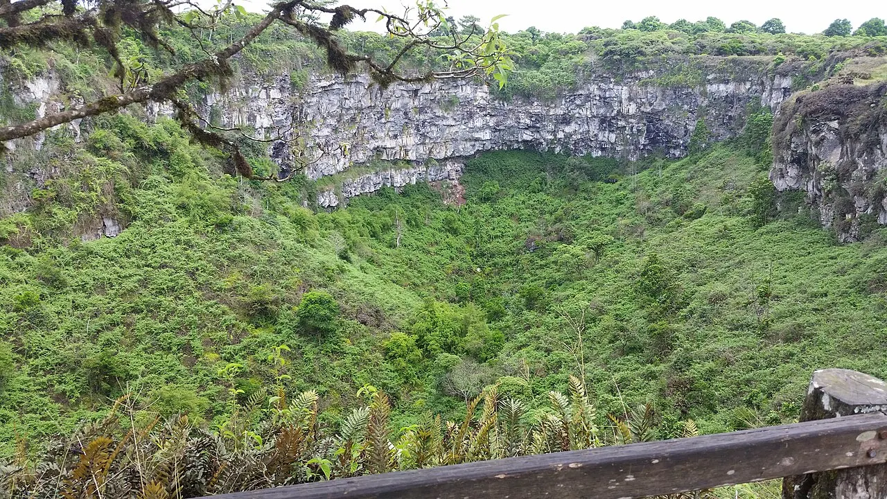 A cratera desce coberta de vegetação até um paredão de rocha, com um corrimão de madeira no primeiro plano.