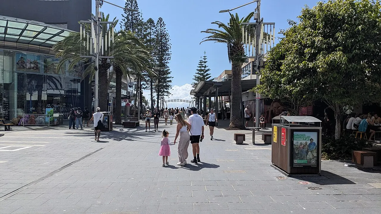 Paved pedestrian street with pines and palms, shops on the left and people walking away towards an arch in the distance.
