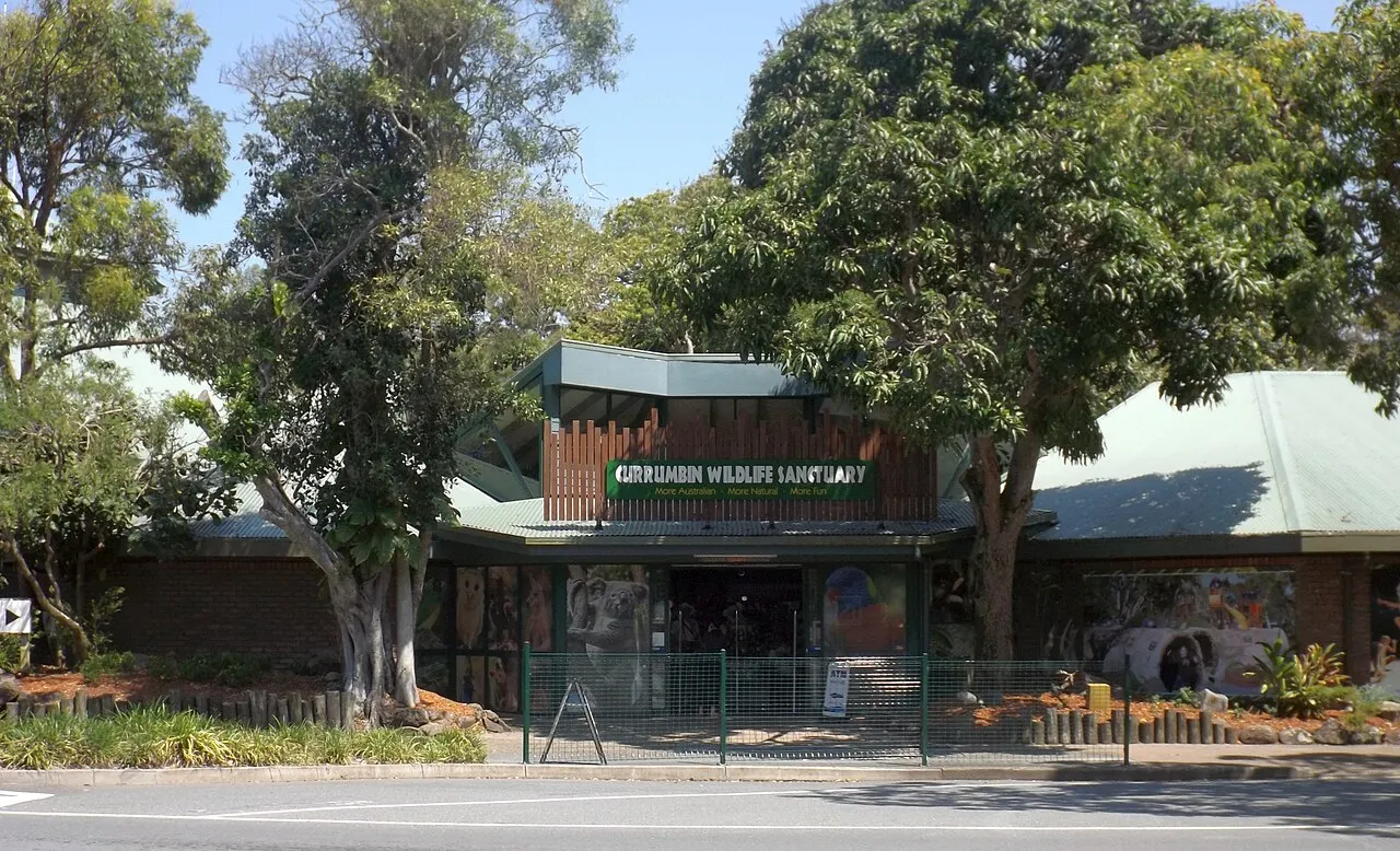Low entrance building with the sanctuary's sign on the facade, among large trees, behind a fence and a road.