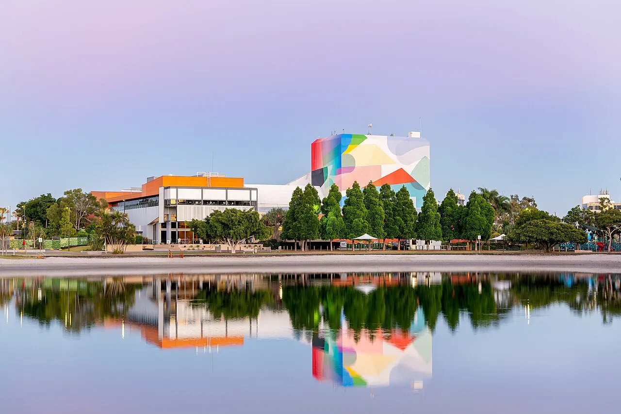 Tall building with a colourful geometric facade beside a low orange and white building, mirrored in still water under a pink sky.