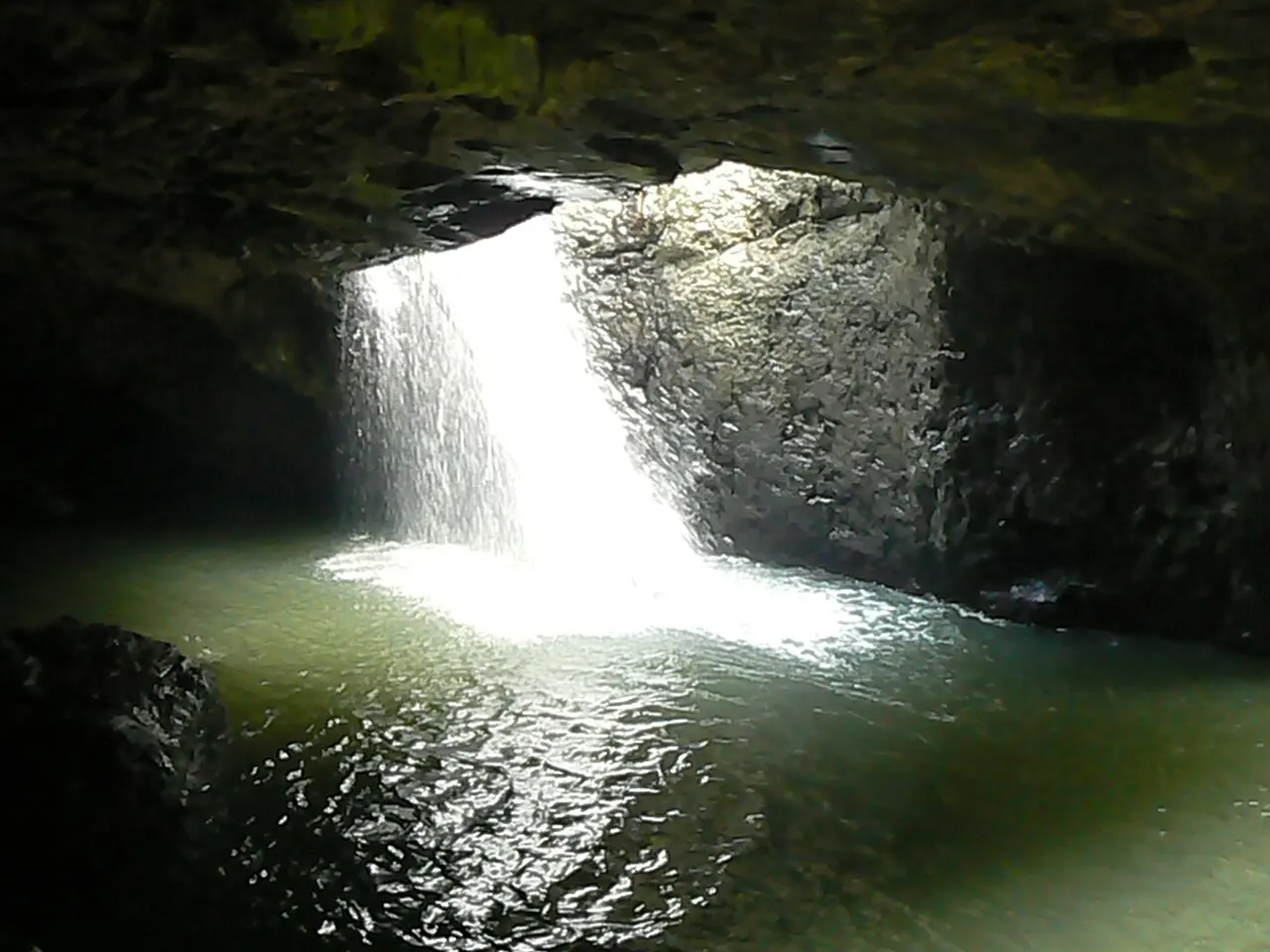 Intérieur sombre d'une grotte rocheuse, avec une cascade éclairée qui tombe par une ouverture du plafond dans un bassin d'eau verte.
