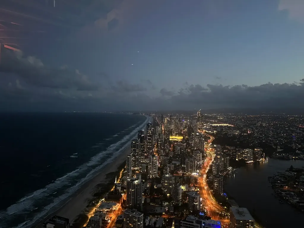 Vue en hauteur, à la tombée de la nuit, de la bande de plage sombre à gauche et des immeubles éclairés le long d'une avenue lumineuse, avec une rivière à droite.