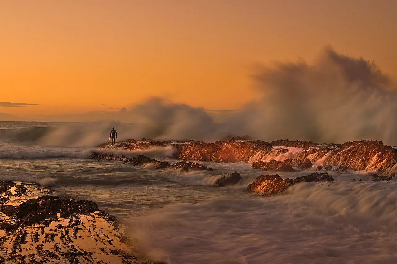 Low rocks at the water's edge lit by a low sun, with waves bursting into spray and one person on the rocks, under an orange sky.