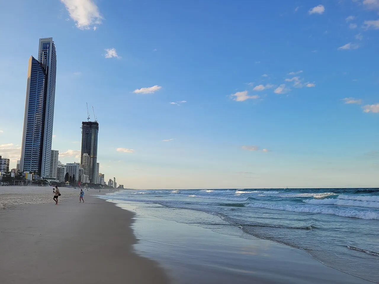 Wide sandy beach with low waves, two people in the distance and high-rise towers on the left under a blue sky with scattered clouds.