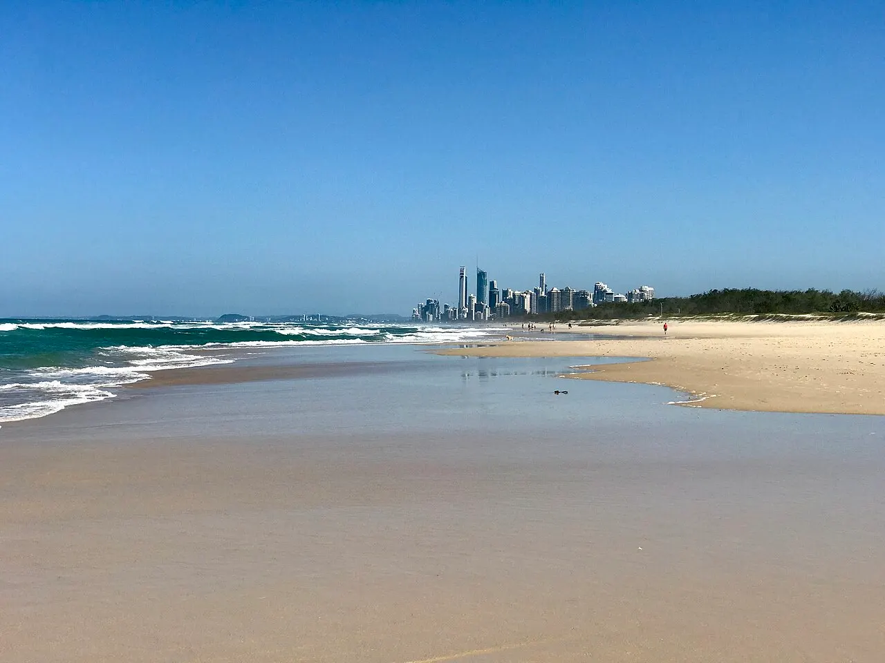 Large plage presque déserte, sable mouillé au premier plan, mer verte à gauche et ligne d'immeubles à l'horizon.