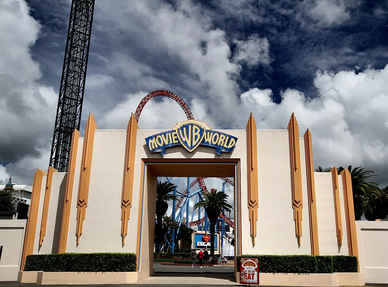 White and gold art deco entrance gate with the park's sign, a ride tower on the left and a roller coaster track behind.
