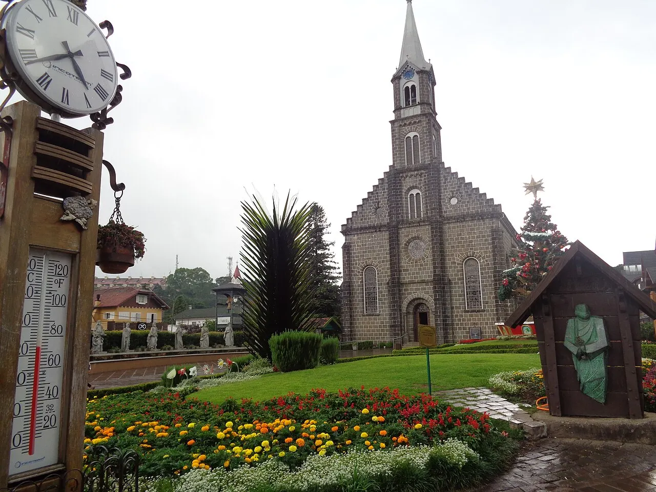 A Igreja Matriz São Pedro, em Gramado, de pedra cinza com uma torre pontiaguda, atrás de um jardim de flores coloridas, com um relógio e um termômetro de rua à esquerda.
