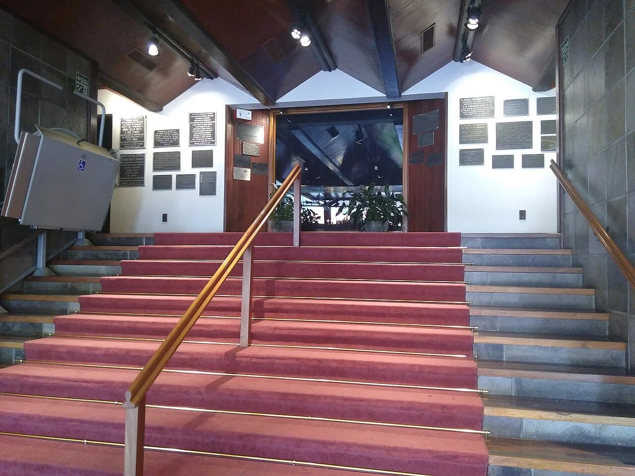 Escadaria interna do Palácio dos Festivais, com passadeira vermelha, corrimãos de madeira, placas nas paredes brancas e teto de madeira.
