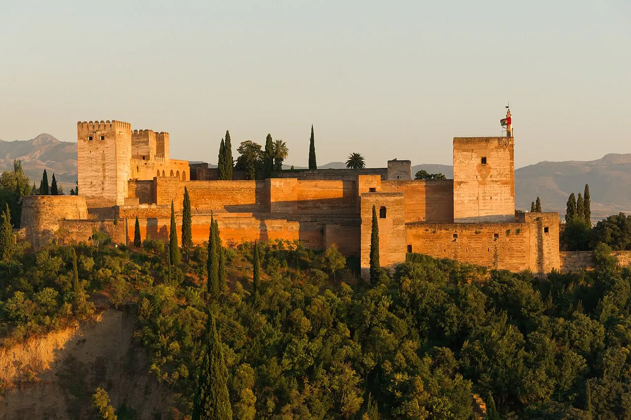 As torres e muralhas da Alcazaba, na Alhambra, iluminadas de laranja pelo sol baixo, acima de um bosque escuro de árvores.