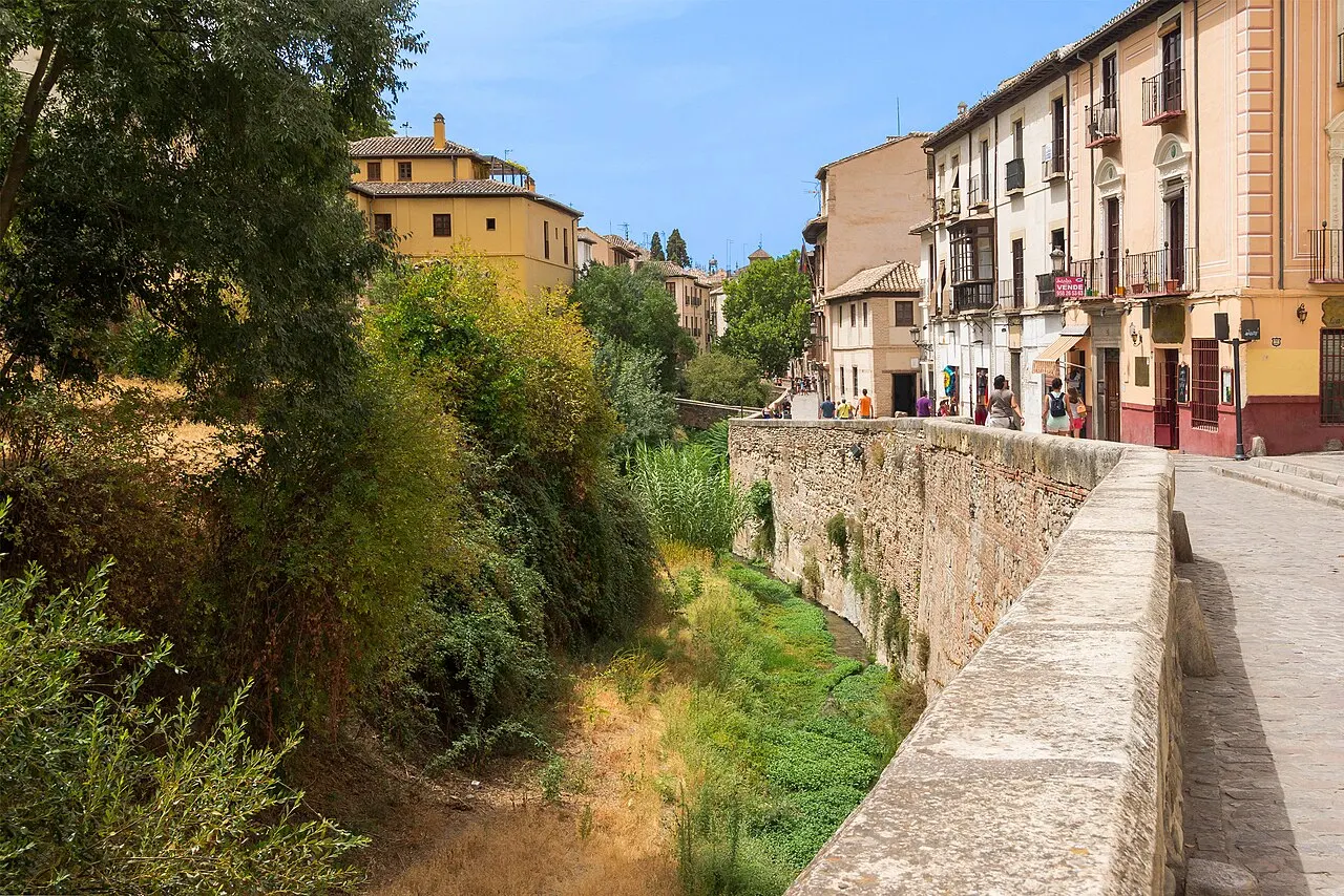 A Carrera del Darro com a mureta de pedra à direita, o leito verde do rio Darro embaixo à esquerda e casas antigas ao fundo.