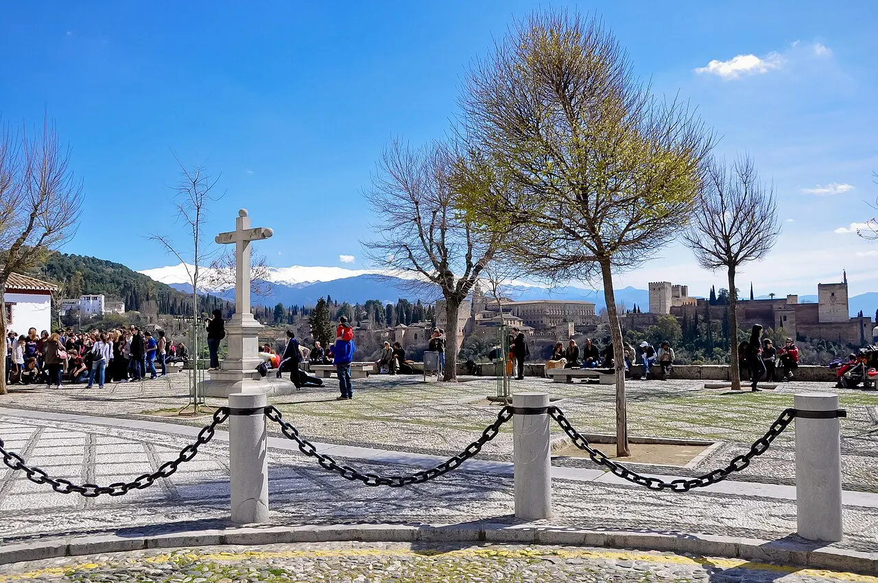 The San Nicolás viewpoint square with a stone cross, chains and bare trees, people at the wall and the Alhambra and snowy Sierra Nevada behind.