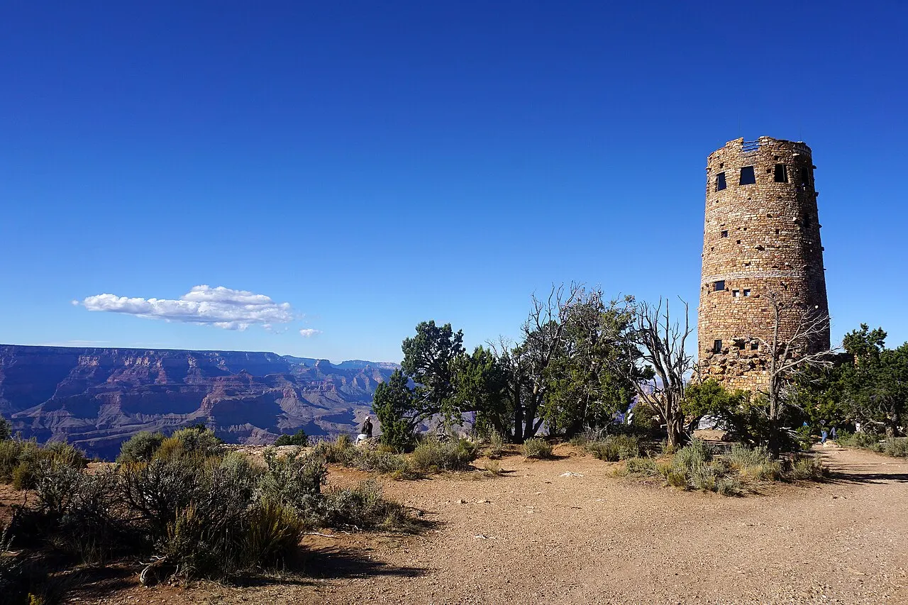 Torre cilíndrica de pedra entre árvores baixas, sobre chão de terra, com o cânion à esquerda sob céu azul.