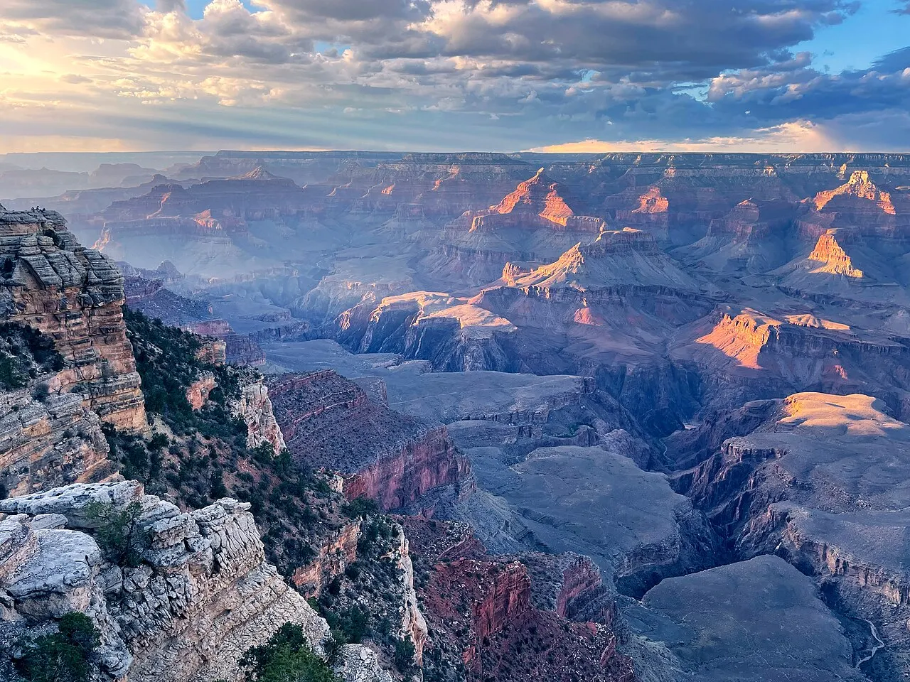 Paredões em camadas do Grand Canyon sob nuvens no fim da tarde, com os topos iluminados em laranja e a borda rochosa à esquerda.