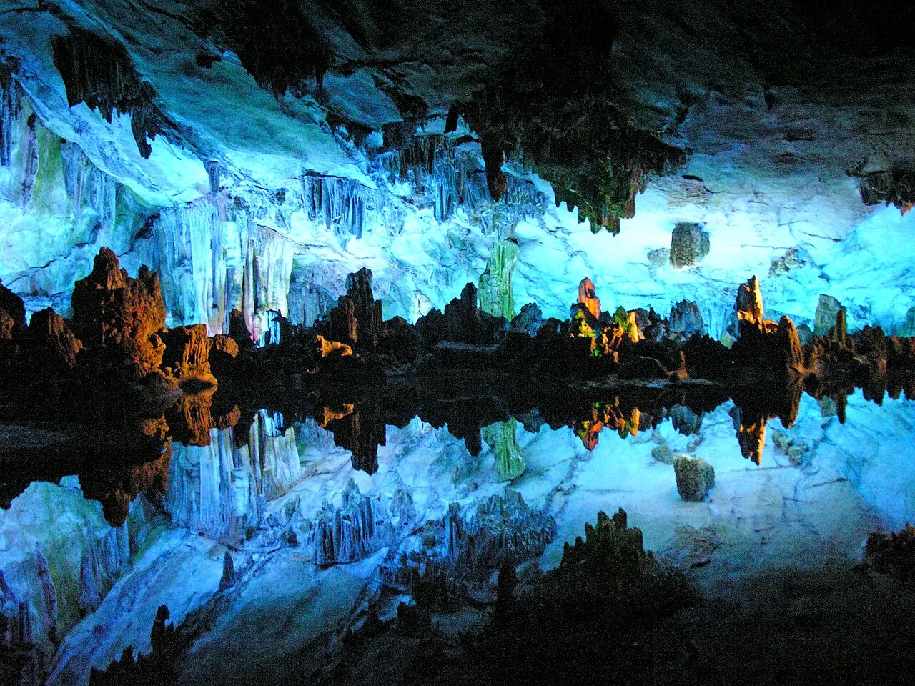 Limestone stalactites and formations lit in blue and orange, reflected in the still water of a pool inside the cave.