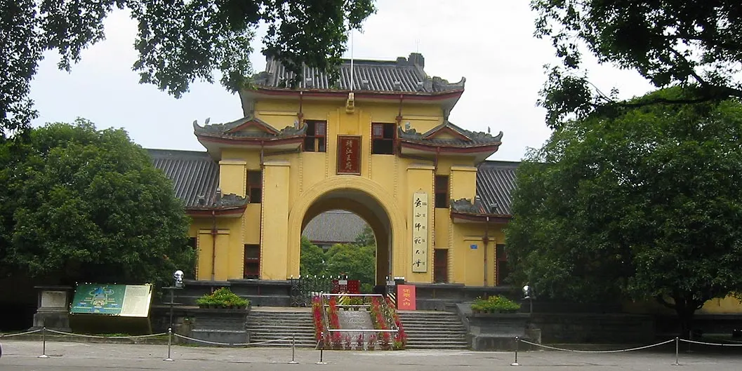 A yellow-walled gate with a central arch and a curved-eave roof, at the top of a flight of steps, between trees.