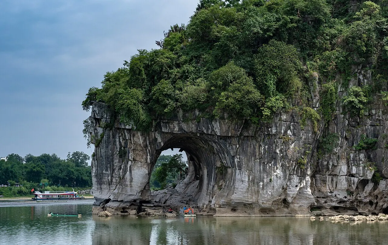The grey rock hill topped with vegetation, with a natural arch open over the water and a moored boat in the background.
