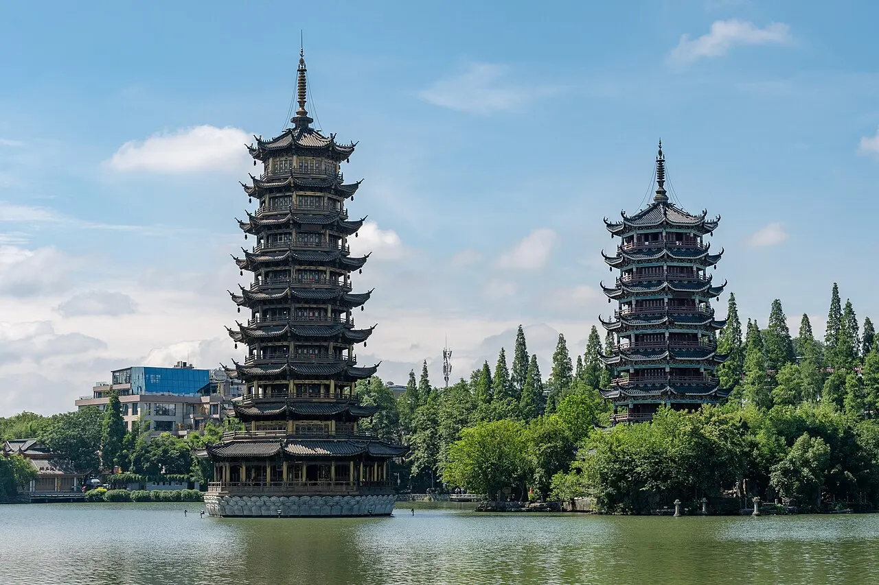Two multi-storey pagodas by a lake, the taller one in front, with trees and buildings behind under a blue sky.