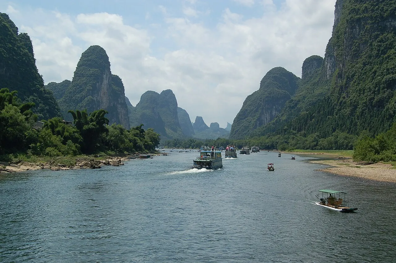 The wide, calm Li River between forested limestone peaks, with a cruise boat in midstream and a smaller raft on the right.