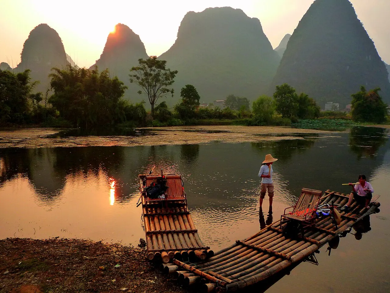 Bamboo rafts moored at the bank at sunset, a boatman in a straw hat standing on one of them and limestone peaks behind.