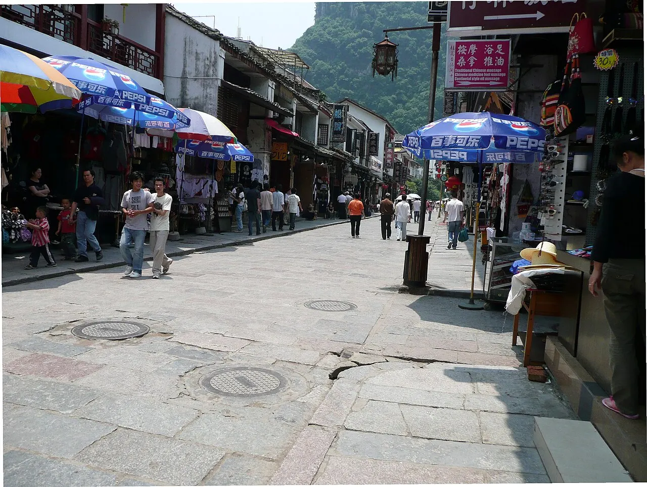 A stone-paved street between shops with blue umbrellas, people walking in the distance and a green hill at the end of the street.