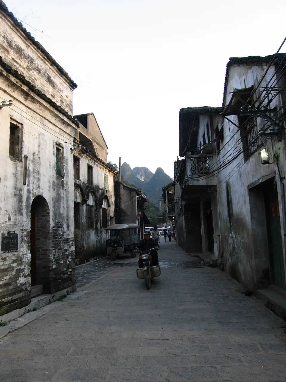 A narrow stone lane between old houses with weathered walls, a motorbike approaching and limestone peaks in the distance.