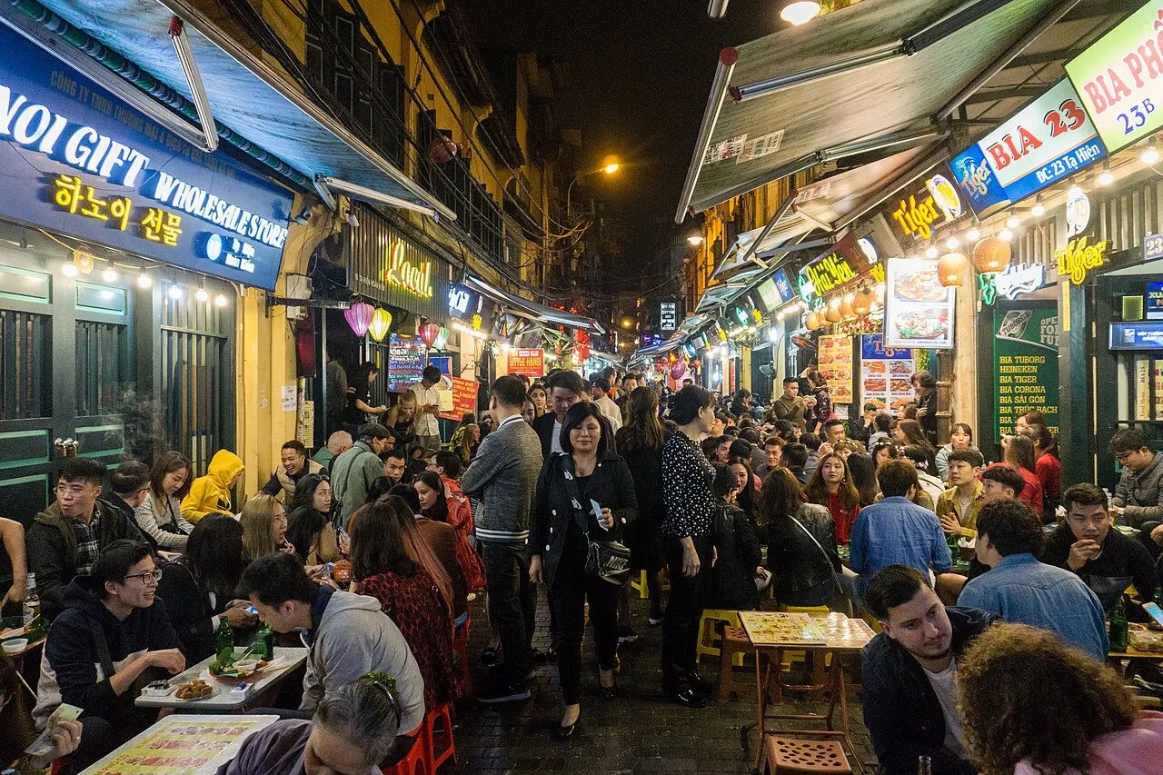 Uma rua estreita à noite, lotada de pessoas sentadas em mesas e banquinhos baixos do lado de fora de lojas e bares, com toldos, lanternas e letreiros luminosos coloridos nas fachadas.