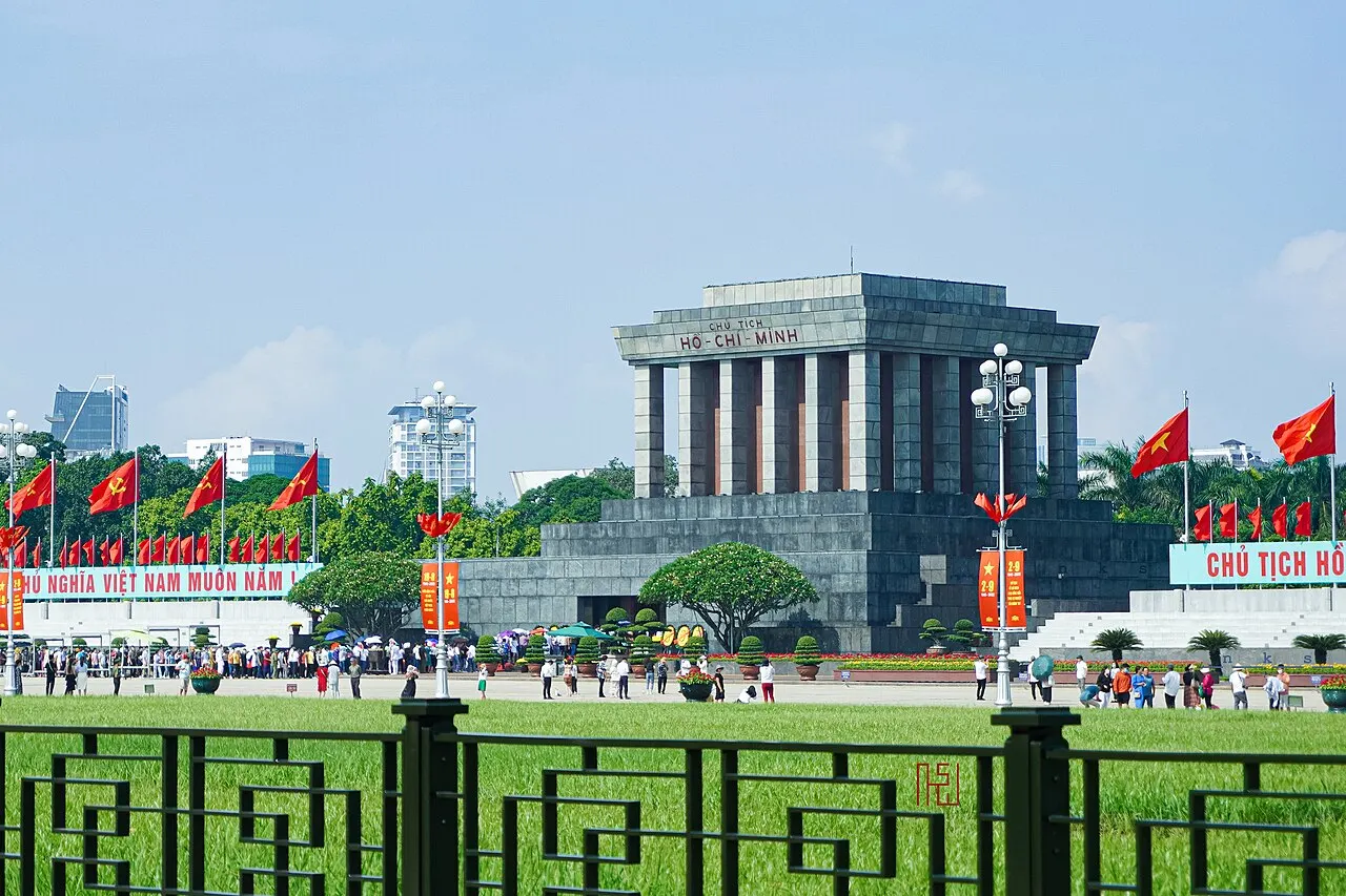 O Mausoléu de Ho Chi Minh, bloco retangular de granito cinza com colunas, elevado sobre base larga, com bandeiras ao redor.