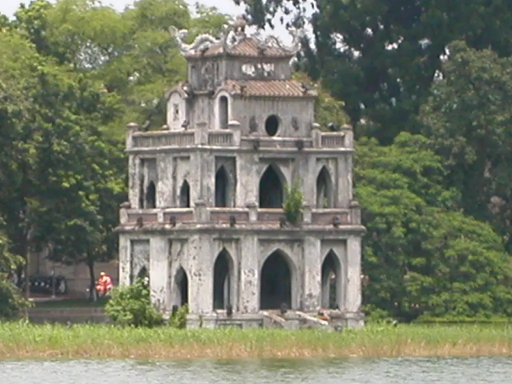 A Torre da Tartaruga, branca e de três andares, sobre um pequeno ilhéu no meio do lago Hoàn Kiếm, com árvores na margem.