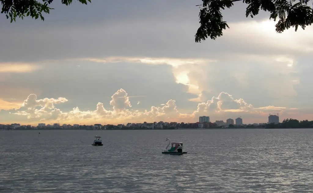 A água ocupa quase o quadro, com dois barcos pequenos, a margem de prédios ao fundo e nuvens no céu.