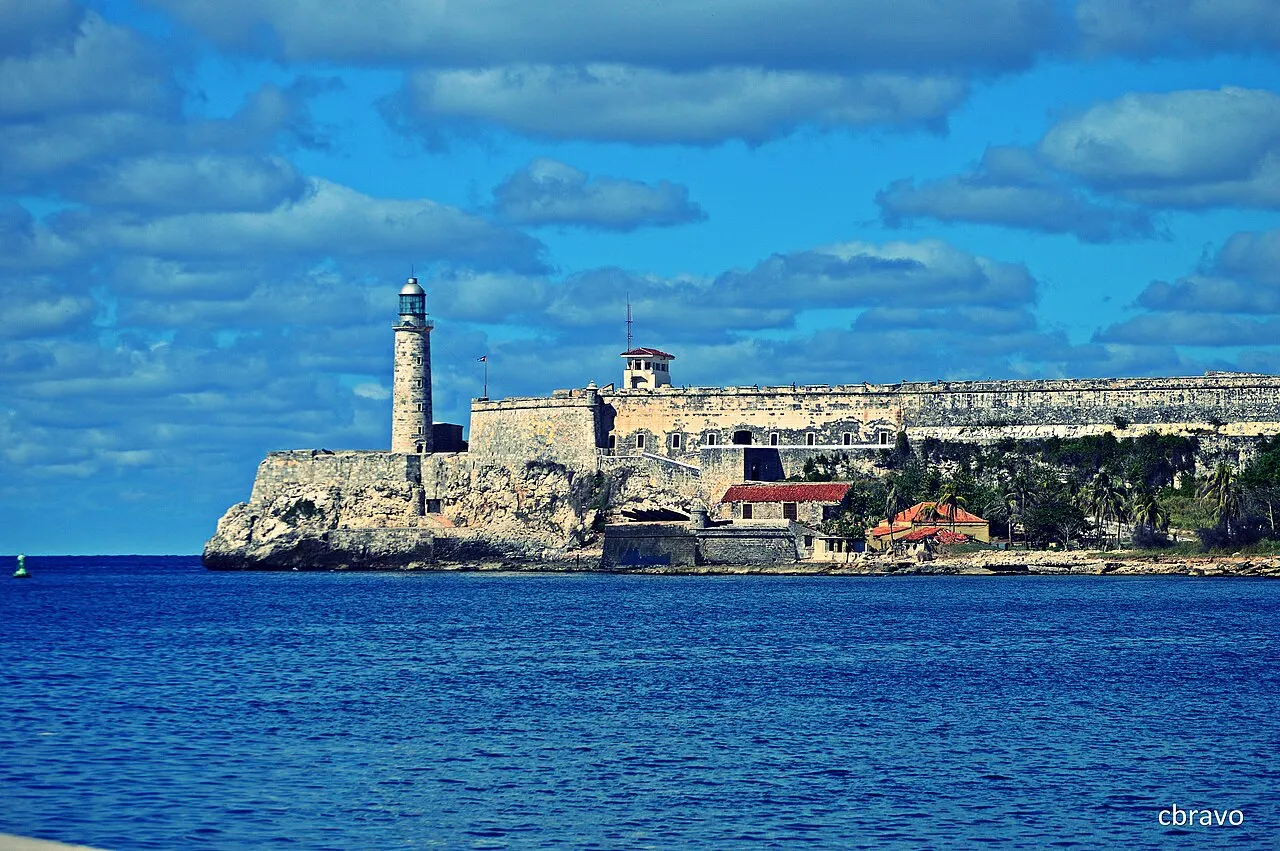 Um farol e muralhas de pedra acompanham a costa sobre o mar azul, com um telhado vermelho junto à água e nuvens no céu.