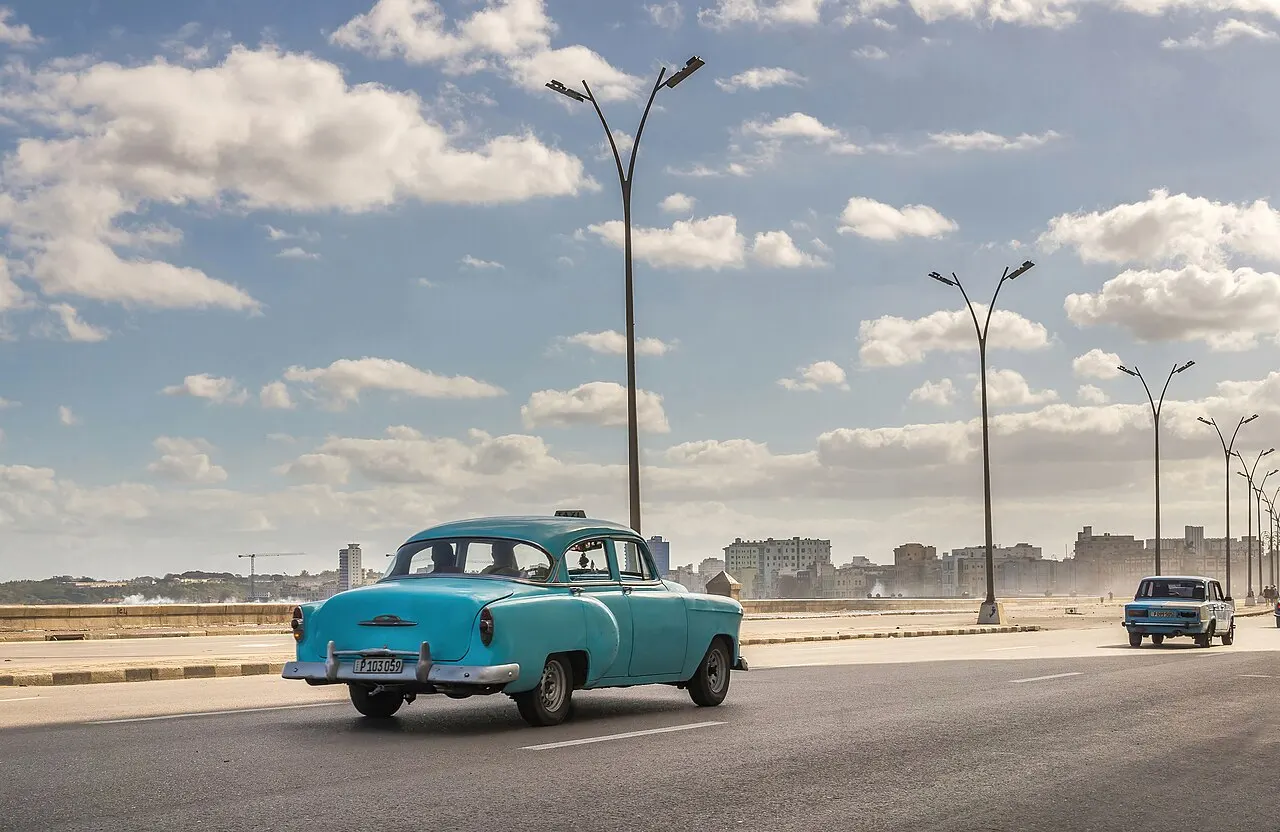 Um carro antigo azul turquesa em primeiro plano numa via larga à beira-mar, com postes de iluminação altos, outro carro ao fundo e prédios da cidade na neblina, sob céu de nuvens esparsas.