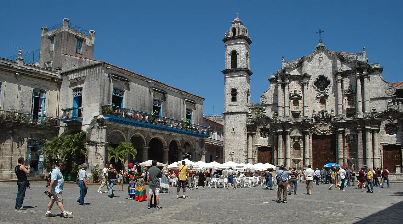 Uma praça de pedra cercada por prédios coloniais desgastados, com a fachada barroca de uma catedral e sua torre de sino num dos lados, mesas brancas sob toldos e pessoas caminhando pela praça, sob céu azul.