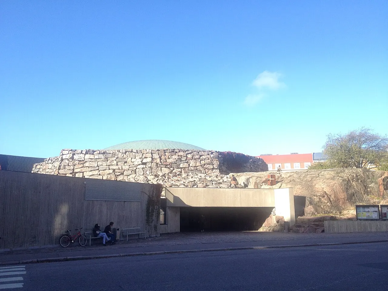 A cúpula de cobre aparece atrás de um morro de pedra irregular, sobre uma entrada de concreto à beira da rua, com duas pessoas e bicicletas num banco.
