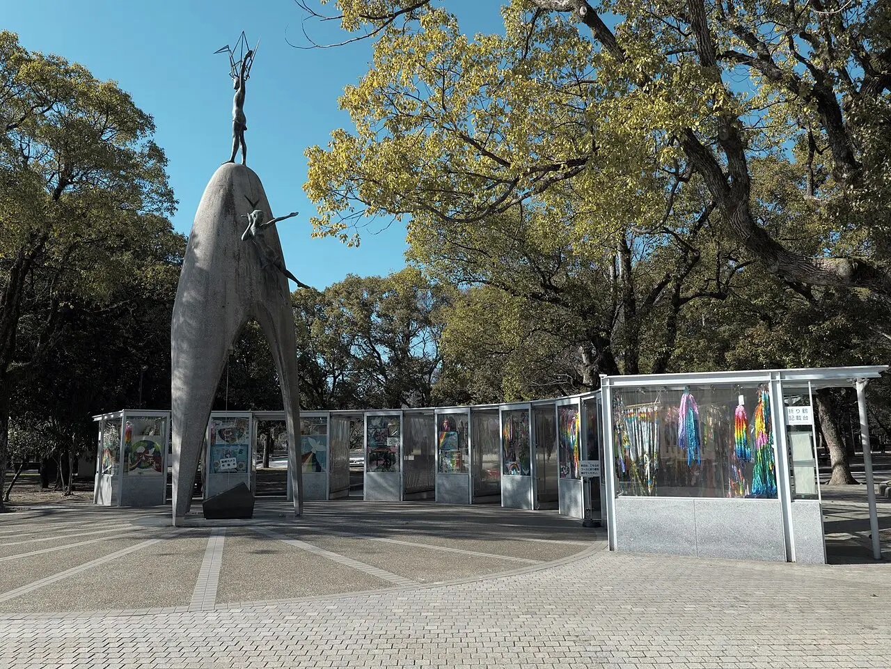 Monumento de pedra com uma figura de braços erguidos no alto, rodeado por vitrines com origamis coloridos e árvores.