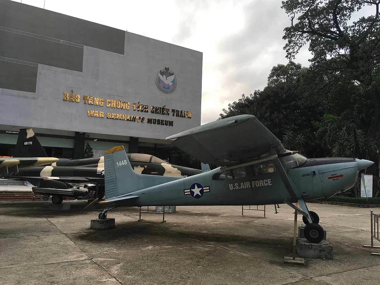 Pequeno avião militar cinza montado sobre suportes, exposto ao ar livre.
