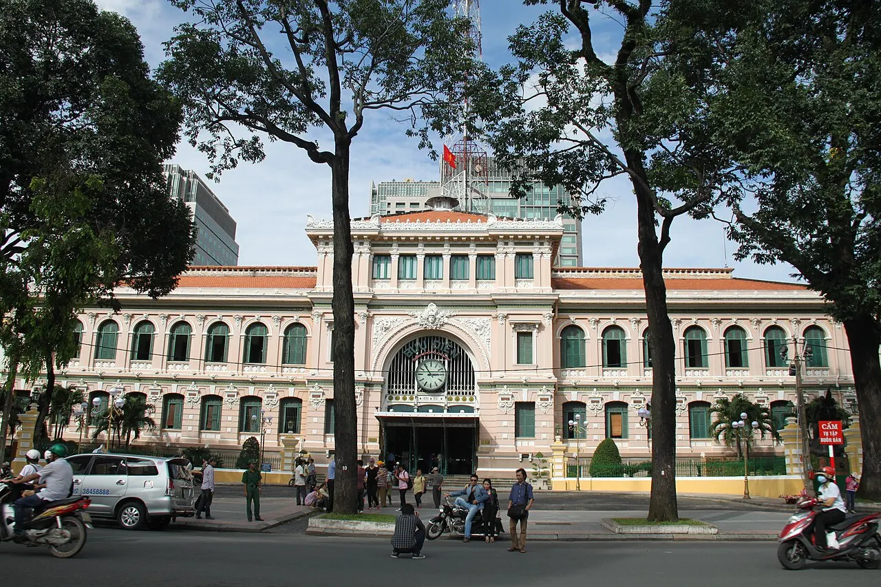 A Agência Central dos Correios de Saigon, prédio colonial amarelo com arcos, relógio central e duas alas simétricas, com árvores em frente.