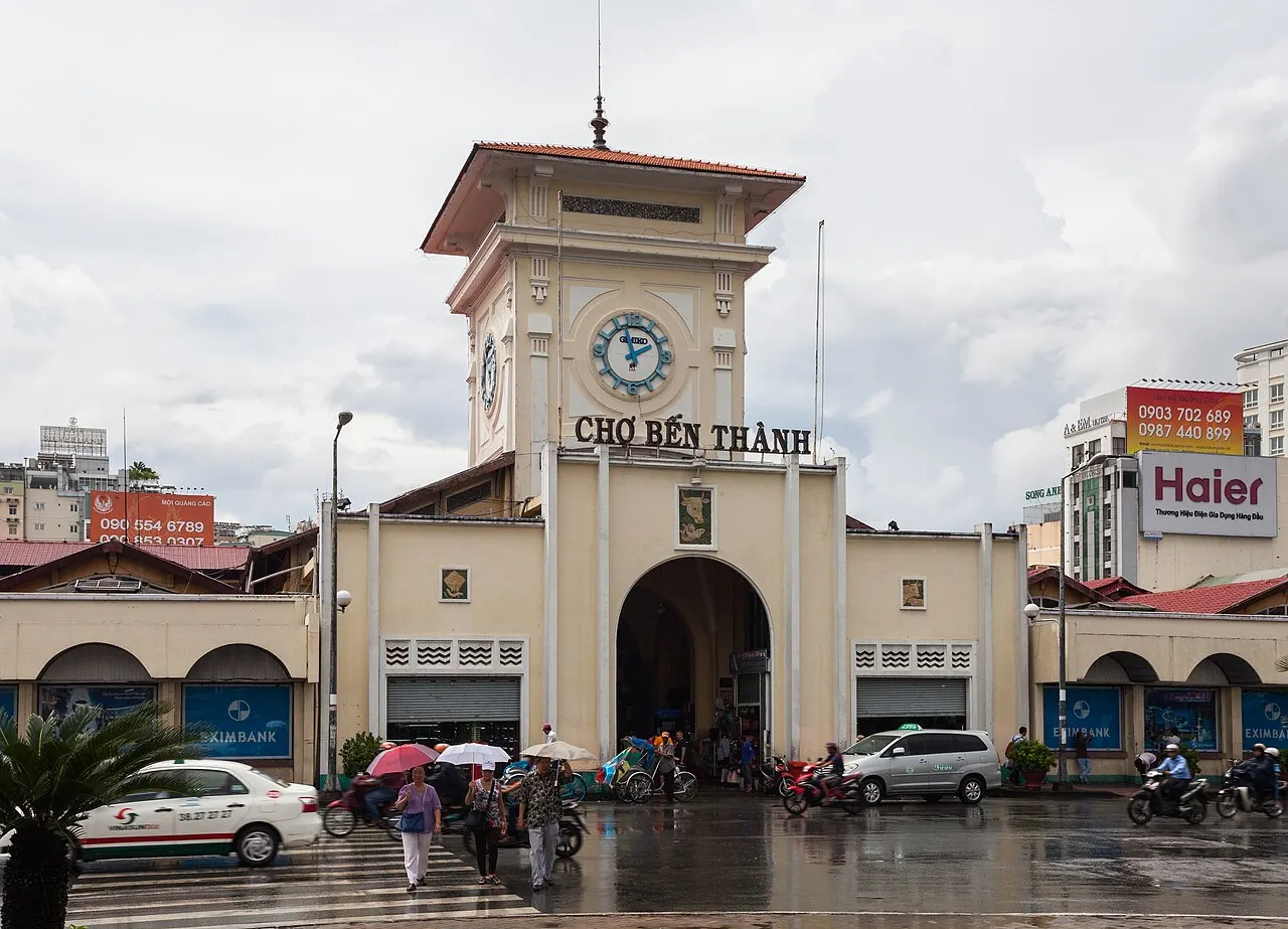 Torre branca com relógio sobre o portão principal do Mercado Bến Thành, com um telhado verde ao fundo.