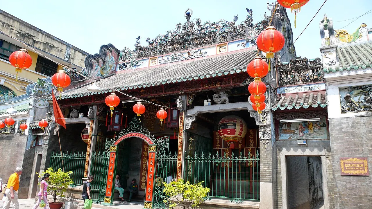 A fachada de um templo chinês, com telhado de cerâmica decorado com figuras esculpidas, lanternas vermelhas penduradas em fileira, um portão de ferro verde na entrada e pessoas passando pela calçada, sob céu claro.