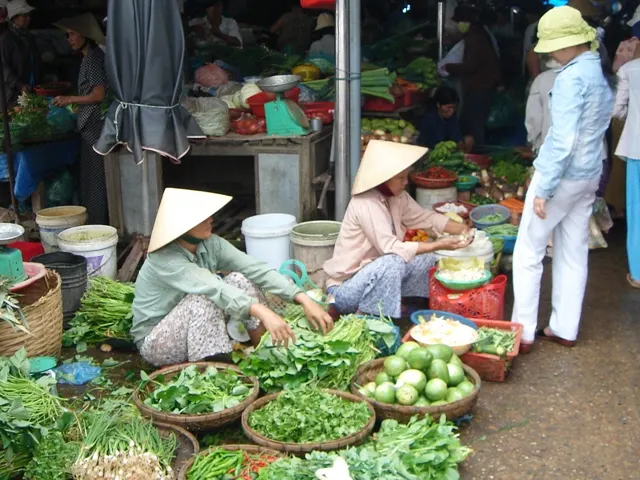 Des vendeuses en chapeau conique assises parmi des paniers d'herbes, de légumes et de citrons verts, avec des étals chargés et des clients debout derrière.