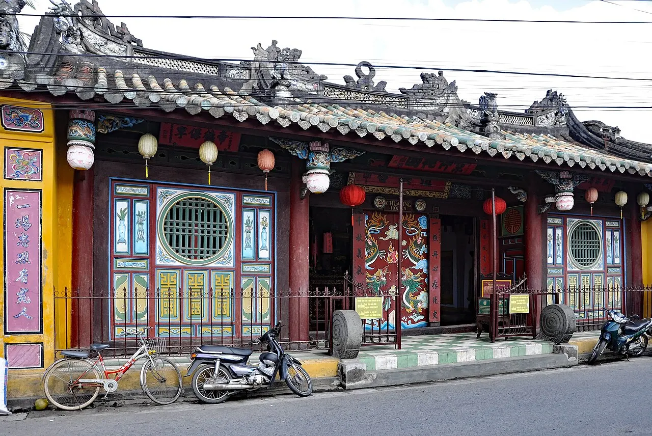 La façade du temple au toit de tuiles orné, aux portes peintes en rouge et vert, aux fenêtres rondes et aux lanternes, avec des vélos et des motos garés sur le trottoir.