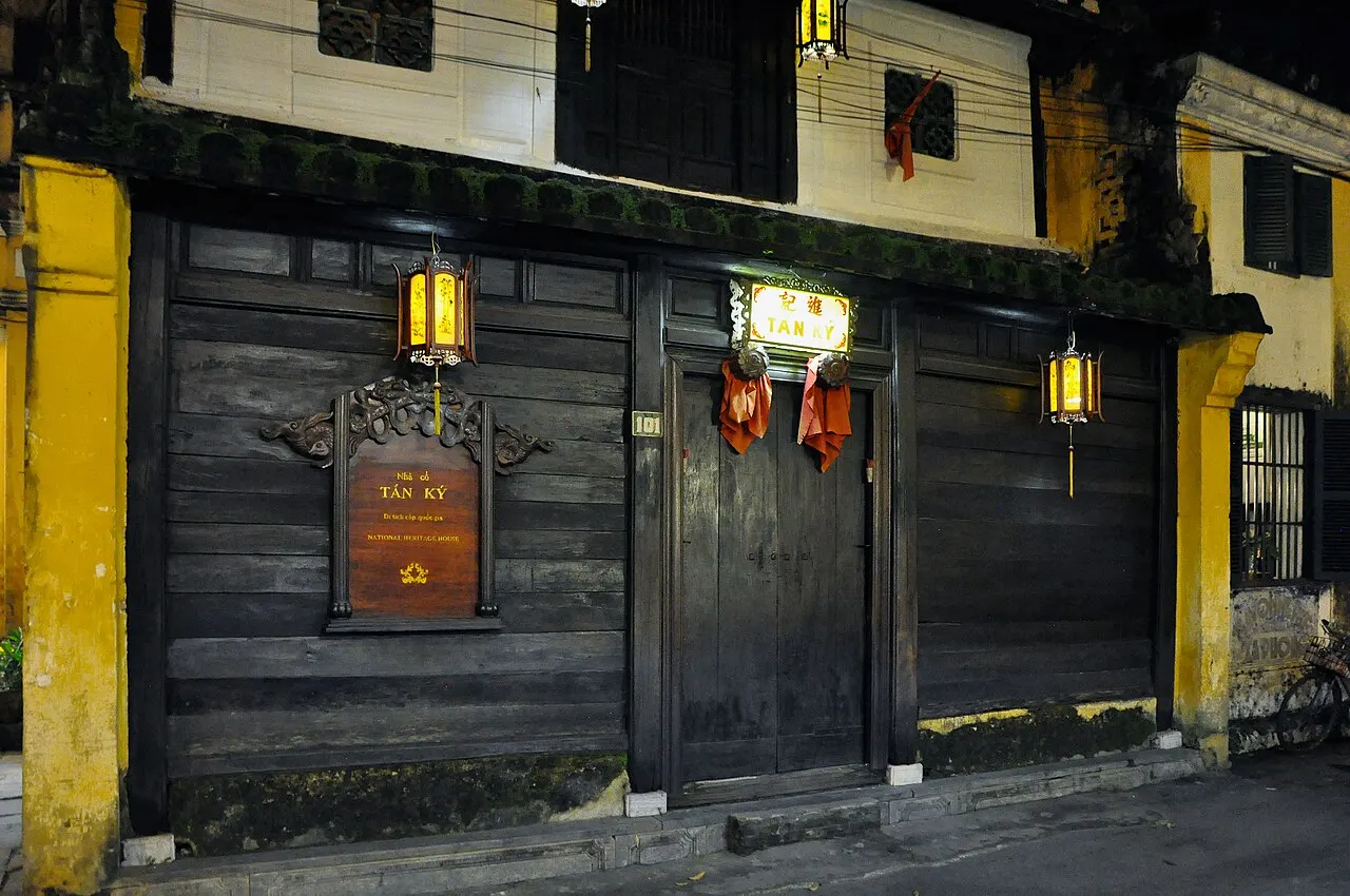 Une façade de planches de bois sombre entre des piliers jaunes, avec des portes fermées, deux lanternes allumées et un panneau sculpté portant le nom de la maison.