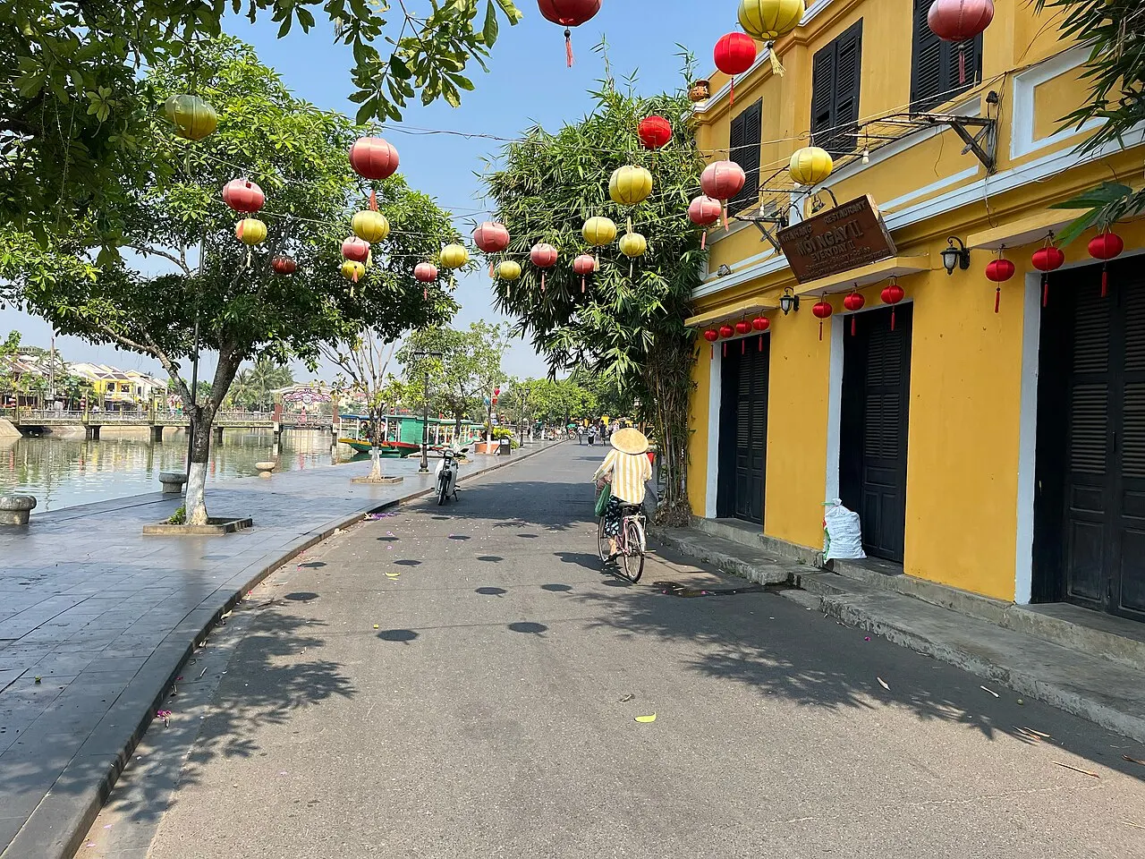 Une large rue au bord de la rivière, avec des maisons jaunes aux volets sombres à droite, des lanternes rouges et jaunes suspendues aux arbres et un cycliste au loin.